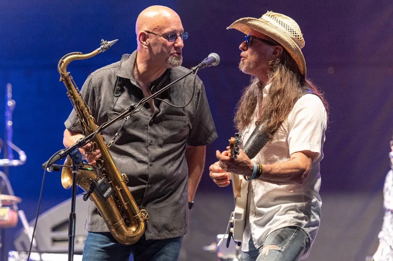 John on saxophone beside a guitarist in a white shirt on stage.
