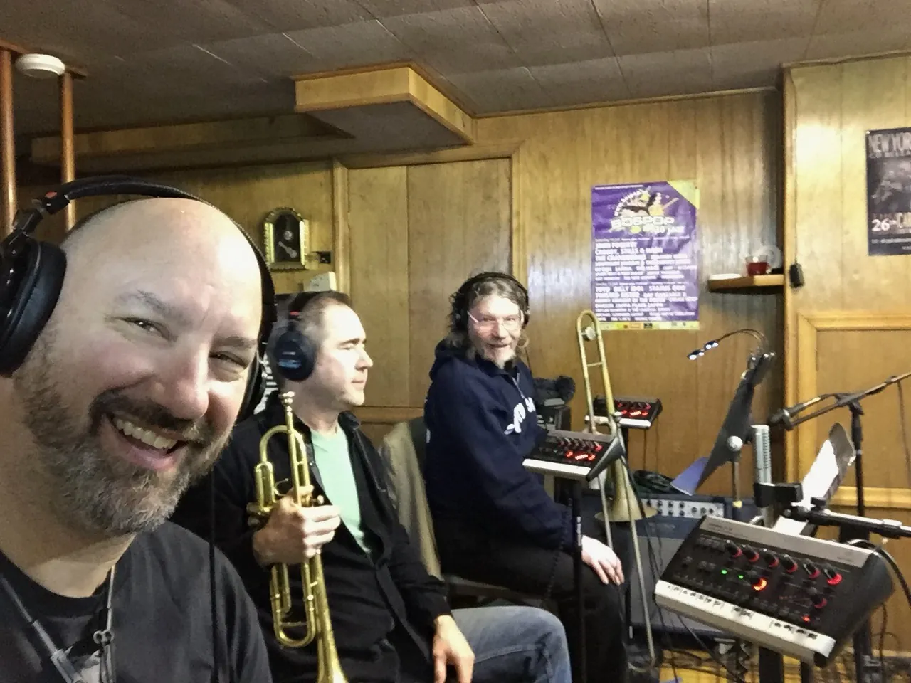 John smiling in headphones beside a trumpet player and a keyboardist in a studio.