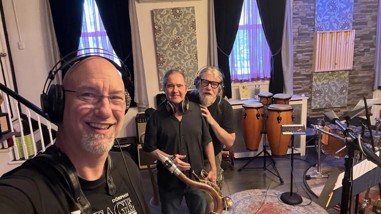 John in headphones in a studio live room, with two musicians behind him.