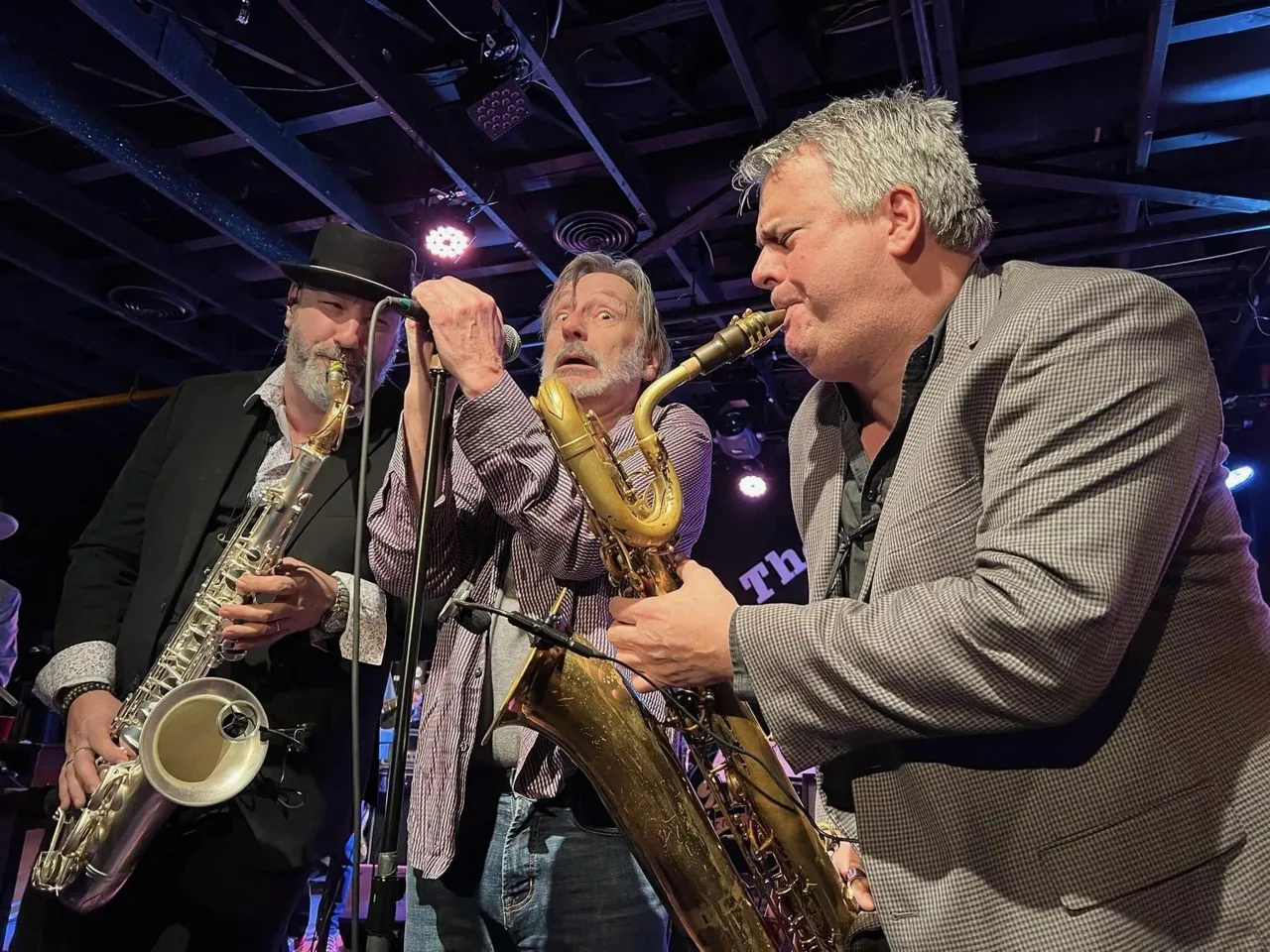 Three saxophonists crowd a microphone on stage.