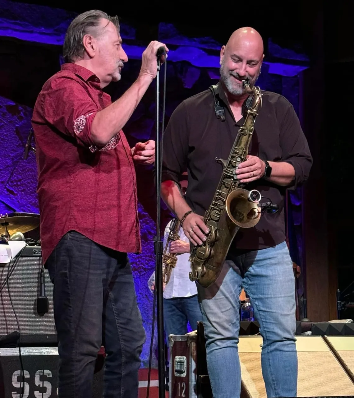 John on saxophone beside a singer in a red shirt on a stone-walled stage.